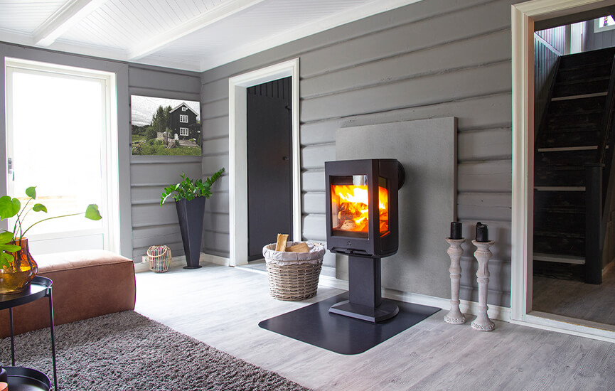 The Jøtul F 167 wood-burning stove in black with glass on three sides in a gray living room with timber walls.