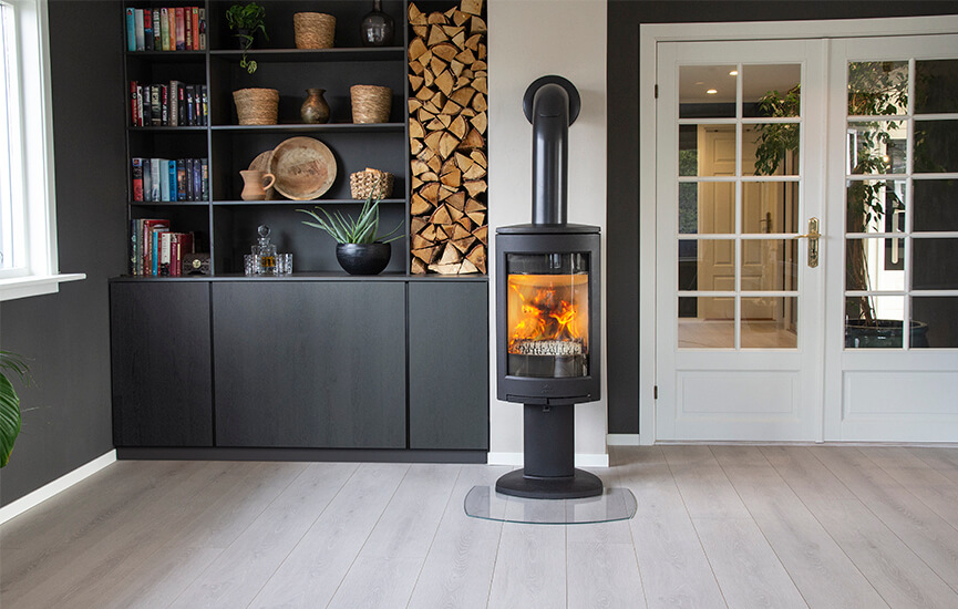 Black wood burning stove Jøtul F 373 Advance in a dark livingroom with a book shelf and firewood niche.
