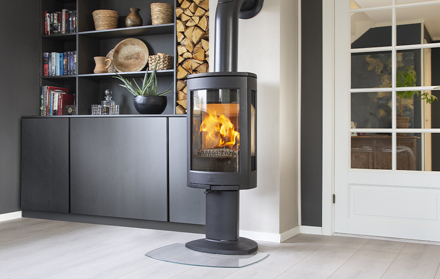 Black wood burning stove Jøtul F 373 Advance in a dark livingroom with a book shelf and firewood niche.